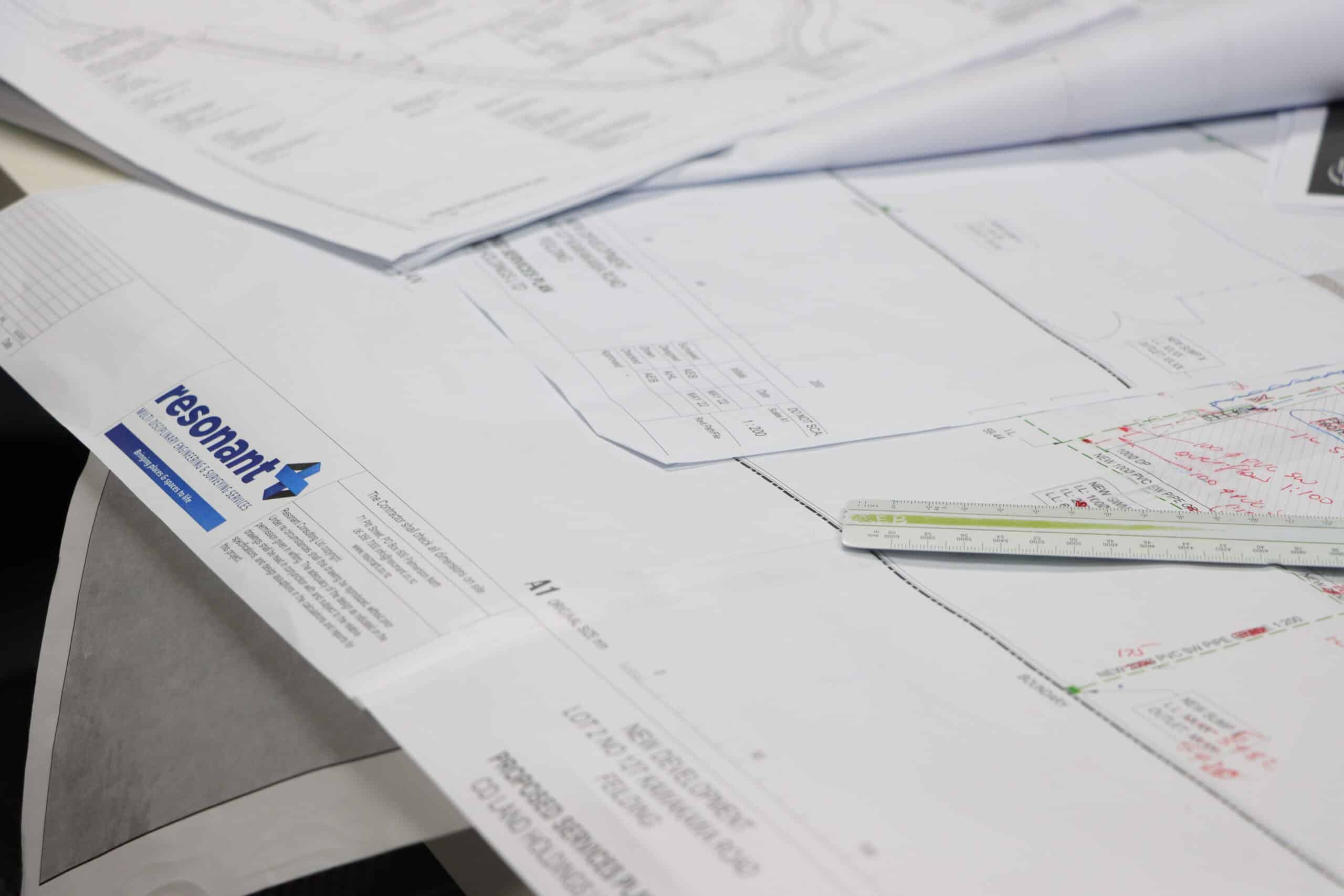 Pile of architectural blueprints and technical drawings spread across a desk with a ruler visible on the right