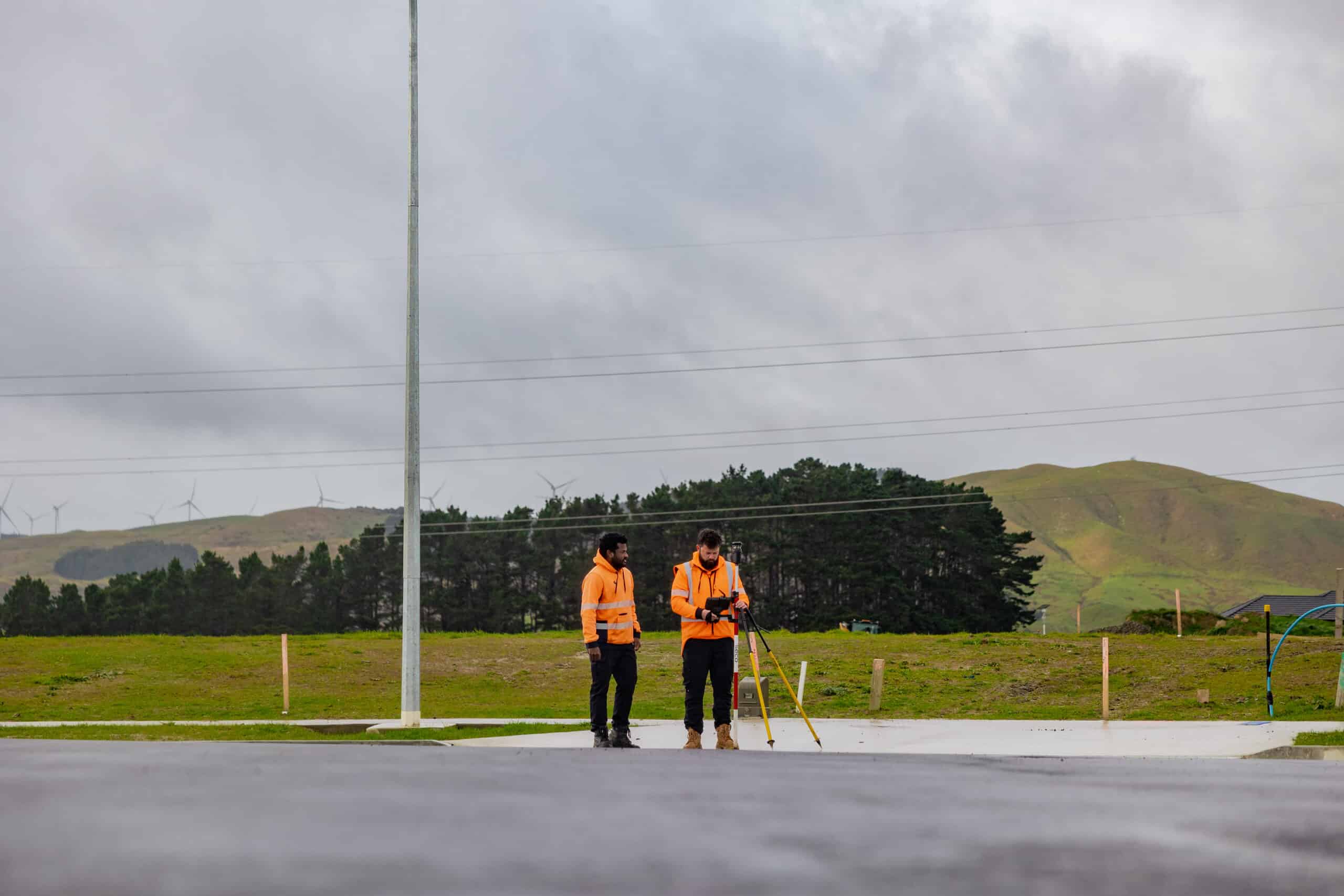 Two workers in high-visibility orange jackets operate surveying equipment outdoors by a power pole, with grassy hills in the background and wind turbines in the distance.