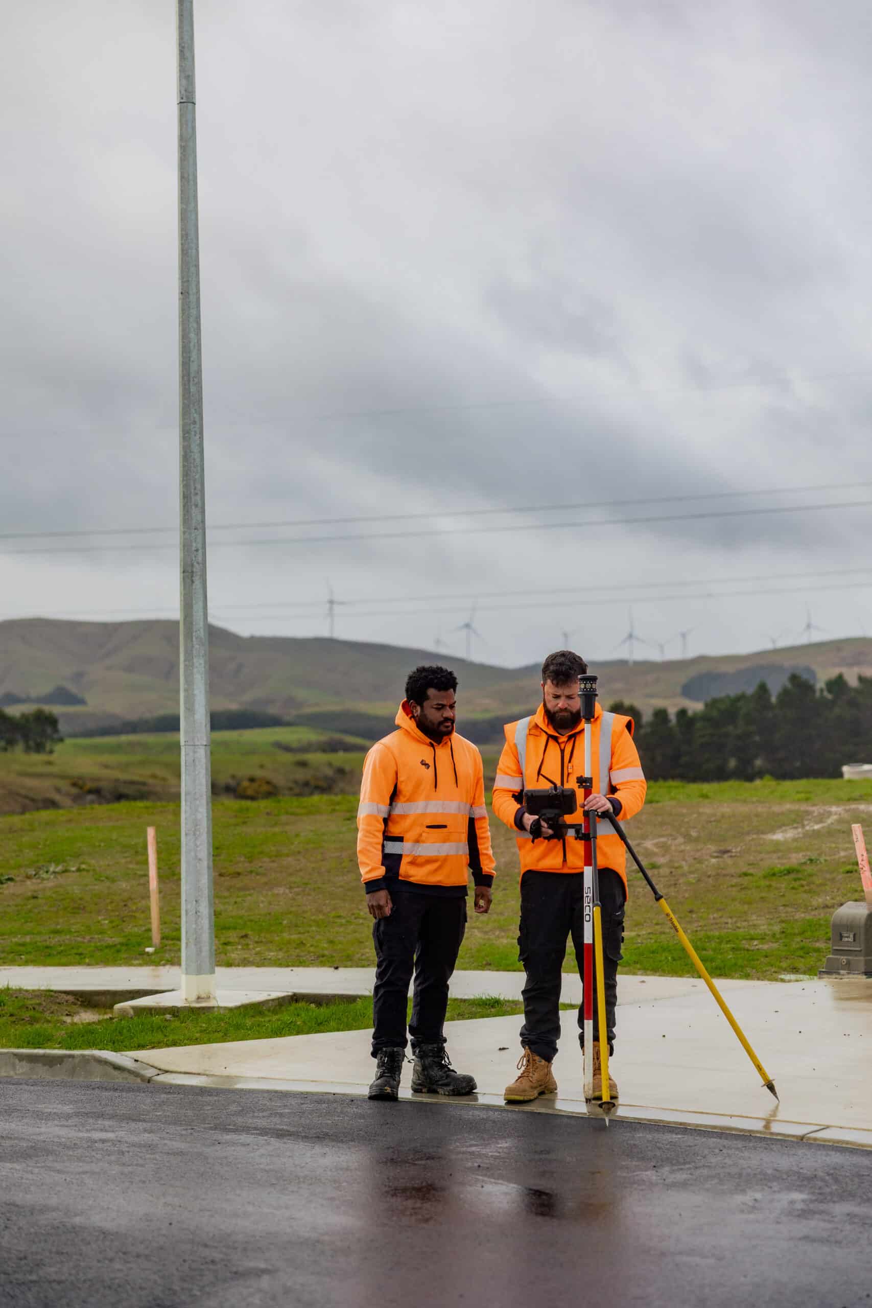 Two construction workers in bright orange safety vests using surveying equipment on a wet road, with wind turbines on hills in the cloudy background.